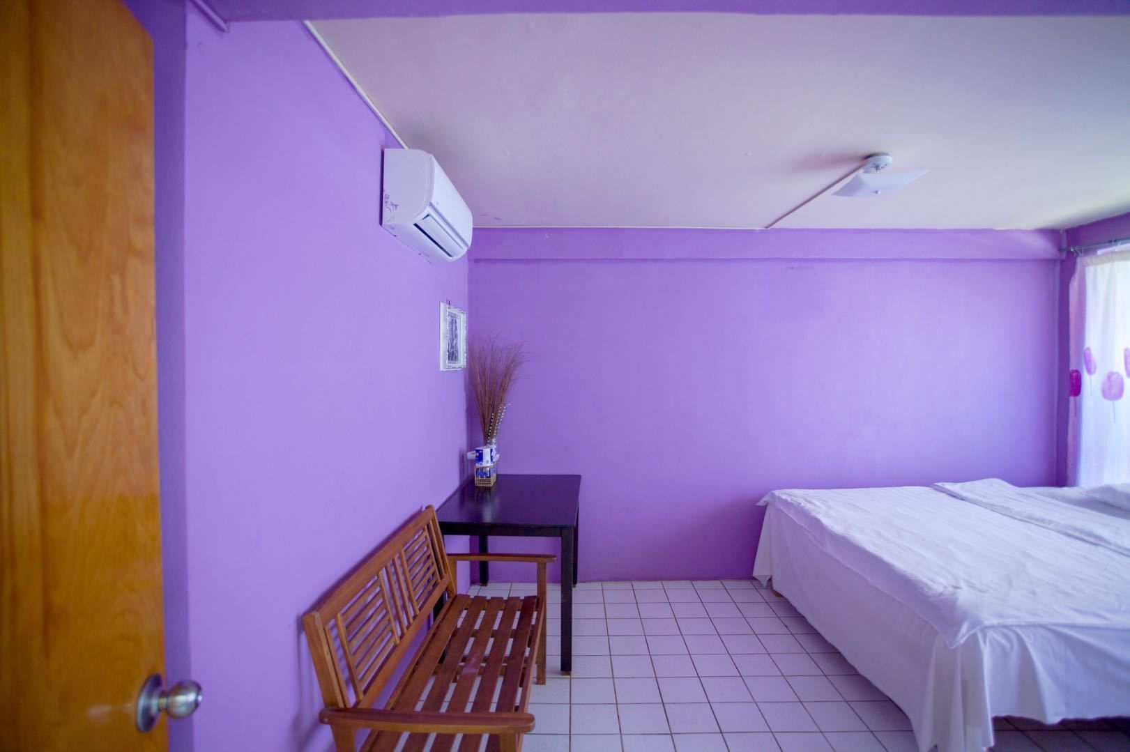 Une pièce aux murs violets, avec un banc en bois, une petite table sur laquelle est posé un vase, un lit et un appareil de climatisation.