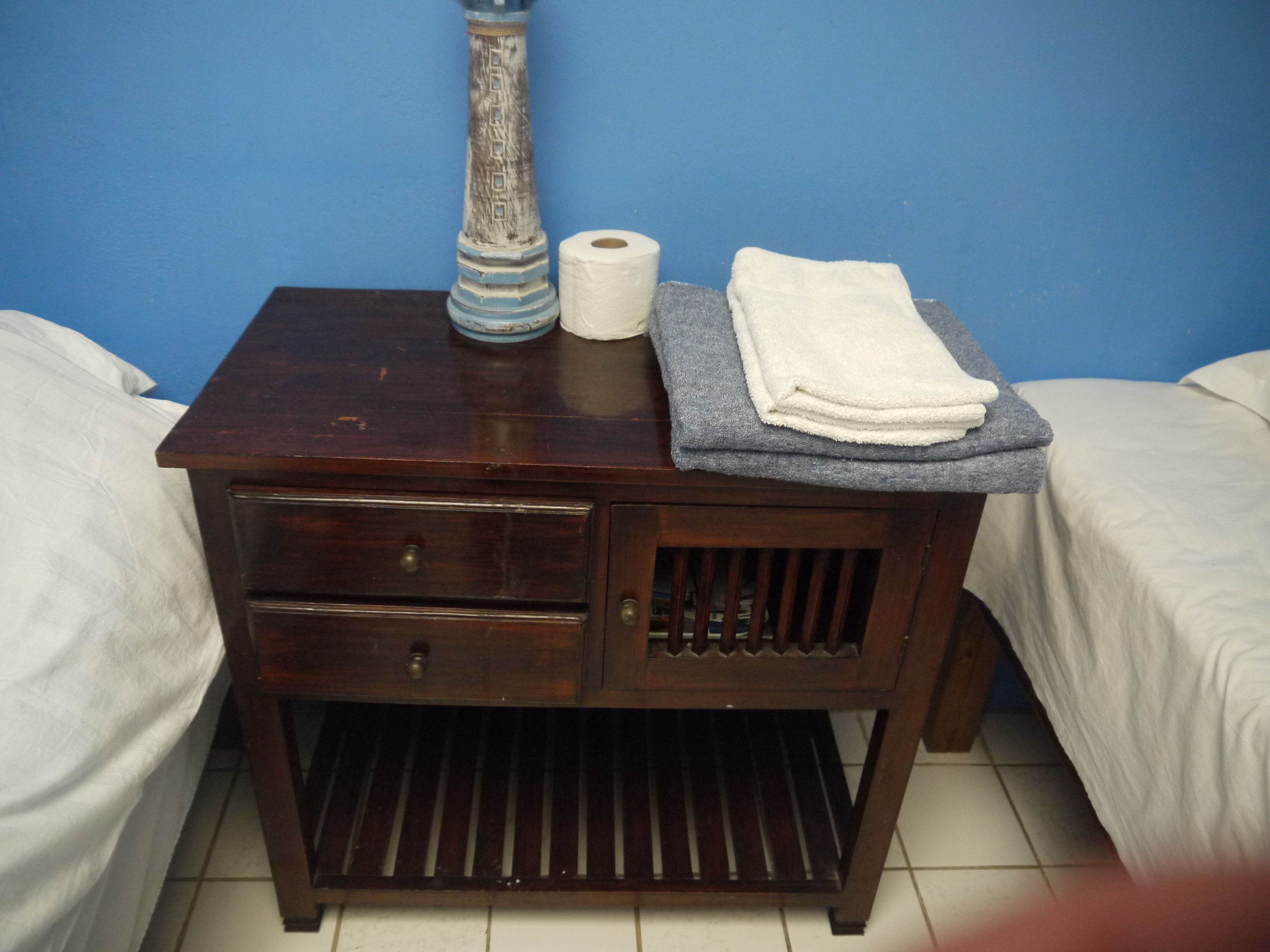 A small wooden bedside table with a vase, a roll of toilet paper, and a folded towel.
