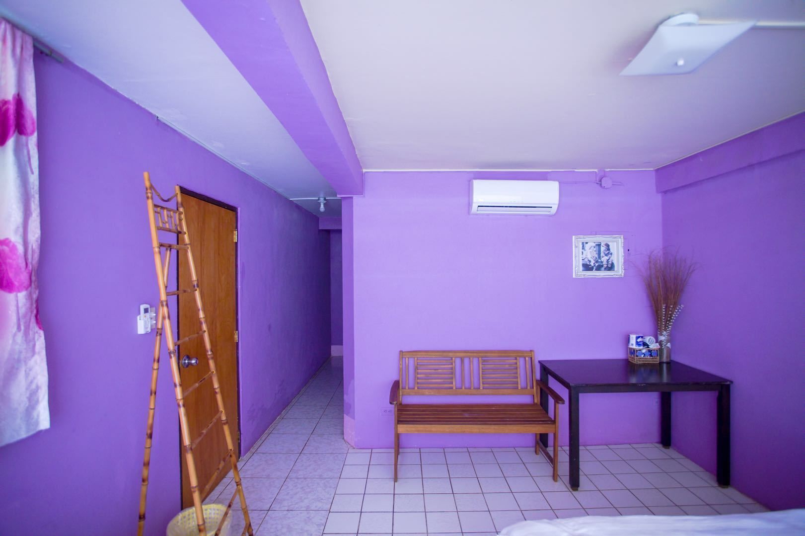 Quarto com paredes roxas, um banco de madeira, uma mesa pequena com um vaso e uma porta com uma escada de bambu.