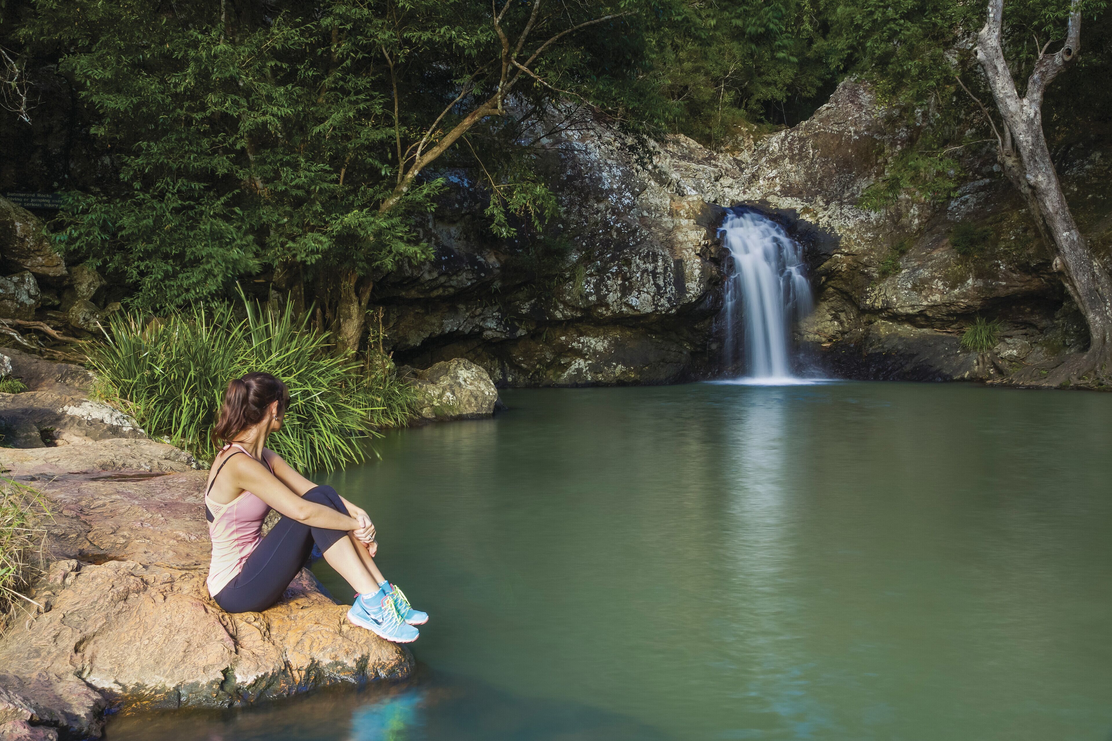 A woman sitting on a rock by a calm waterfall in a forested area.