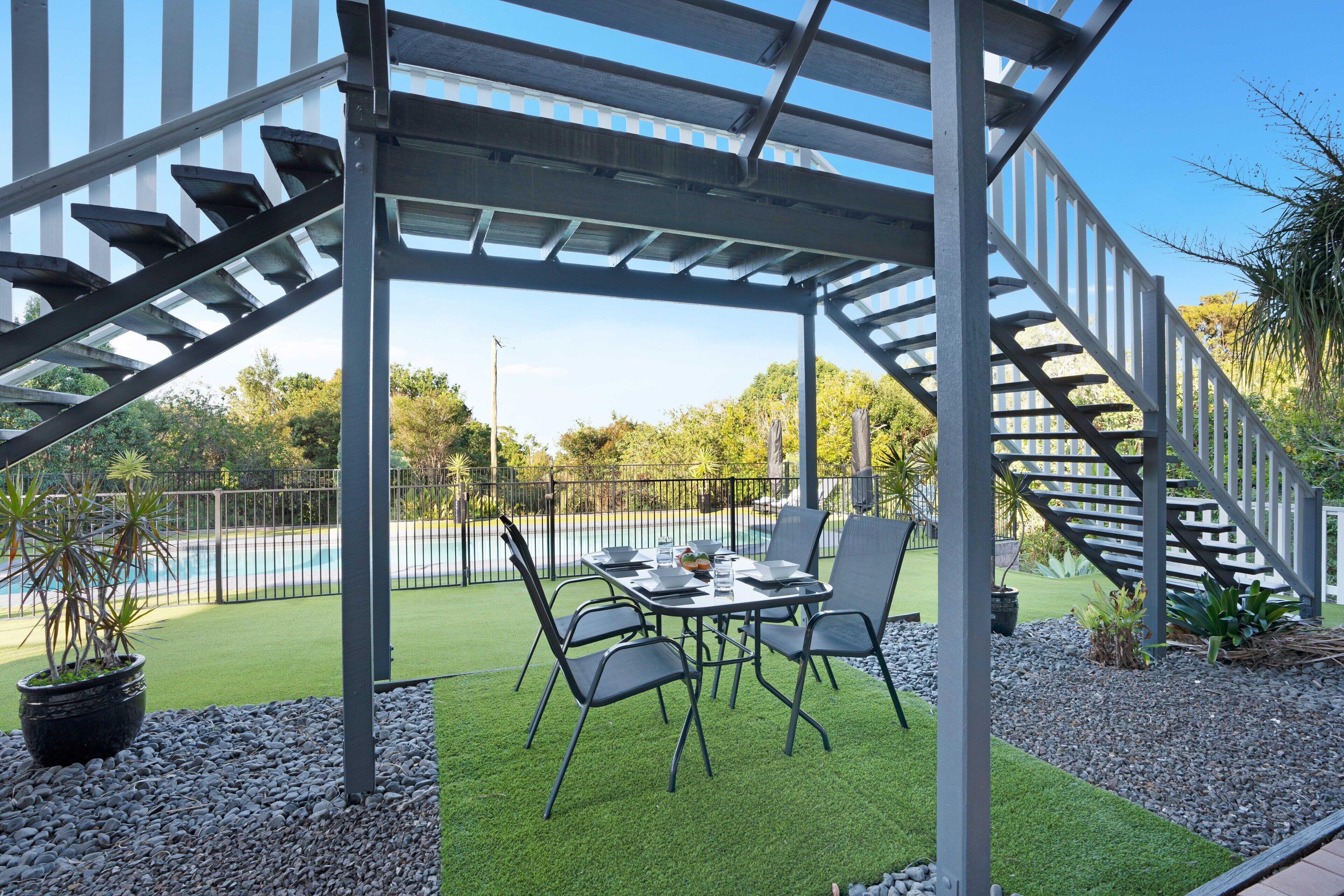 A covered outdoor patio with a table and chairs, surrounded by a pool and greenery.
