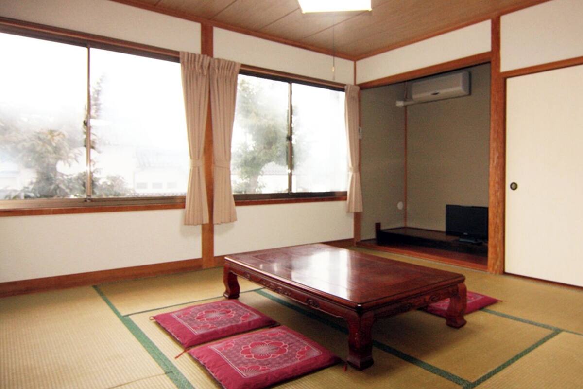A traditional Japanese room with tatami flooring, a low wooden table, sliding doors, and a large window with a view of trees.