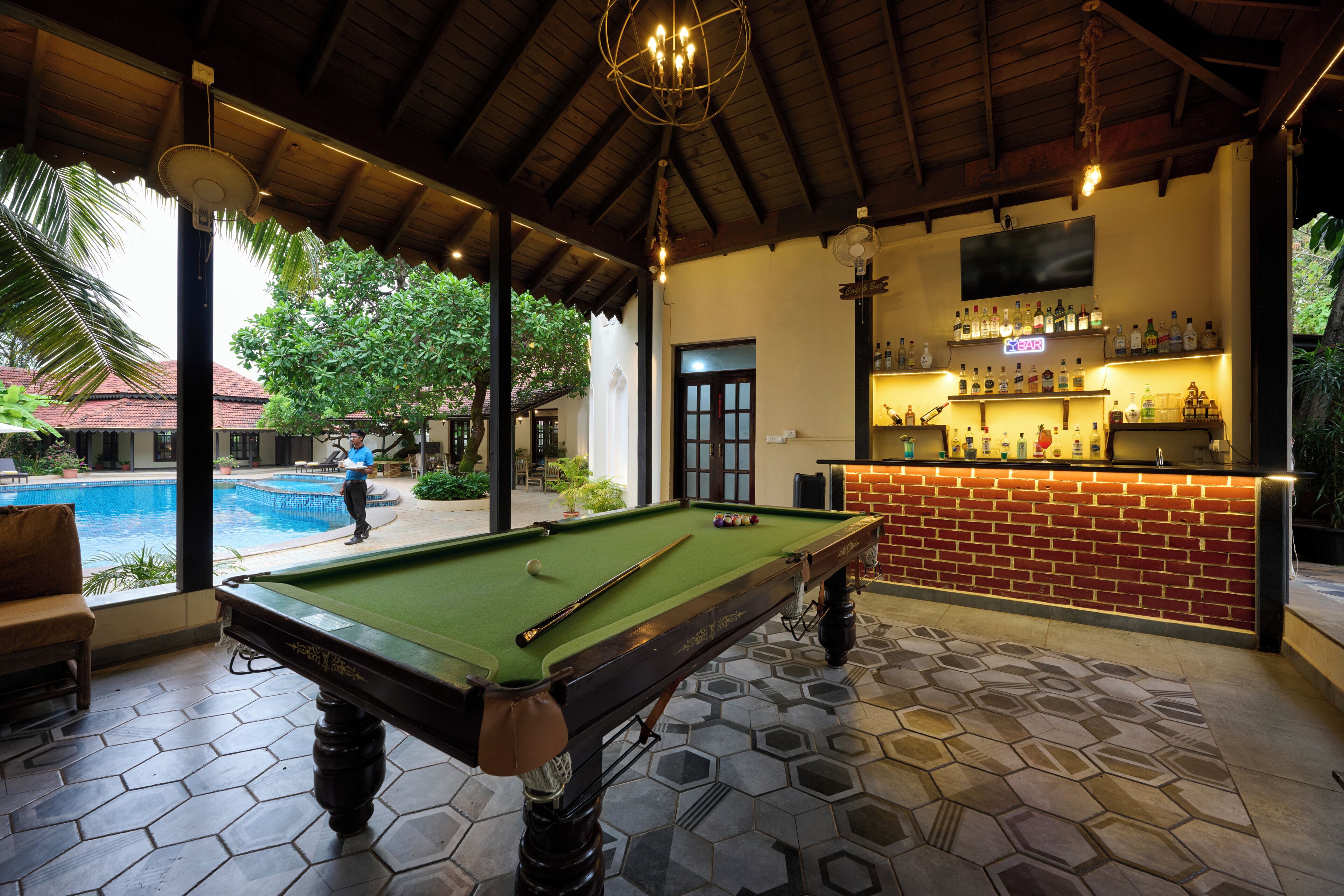 A pool table with a billiard cue and balls, a bar area with bottles, and a pool in the background.