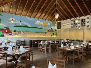 Ein Restaurant mit Holztischen und -stühlen, einem Wandgemälde mit Strandmotiv und hängenden Laternen.