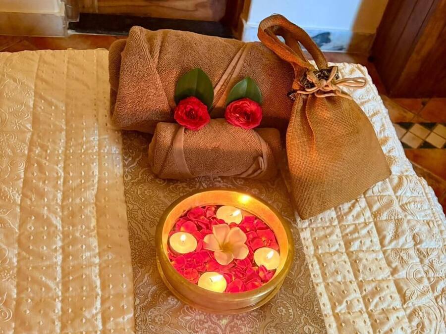 Una cama con colcha blanca, un tazón pequeño de pétalos de rosa y velas, y un arreglo con forma de corazón de pétalos de rosa.