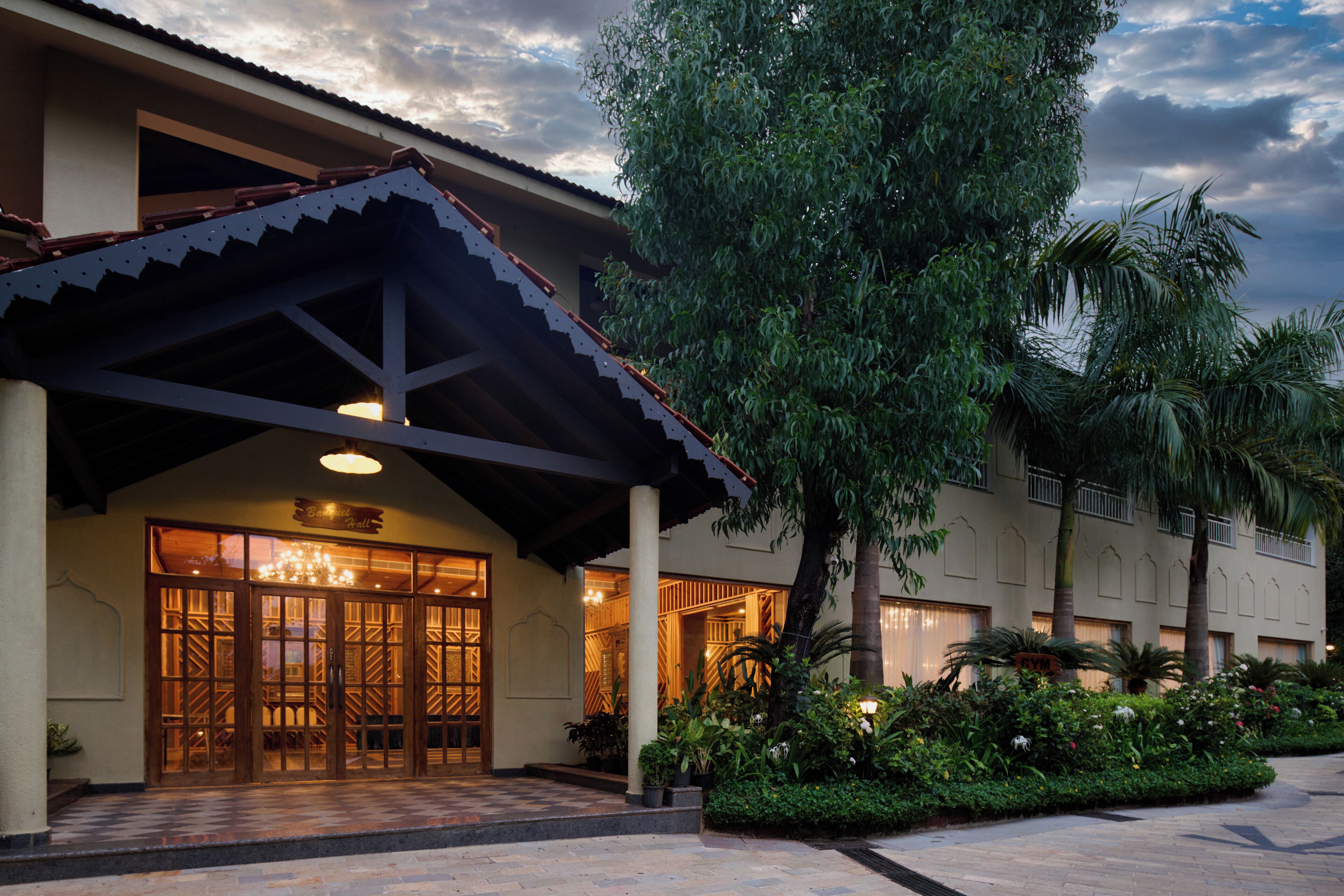 A hotel entrance with a covered patio, large glass doors, and a well-maintained garden with various plants.