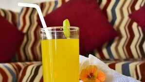 Een glas geel drankje met rietje en een muntblaadje, geplaatst op een tafel met een opgerolde handdoek en een bloem ernaast.