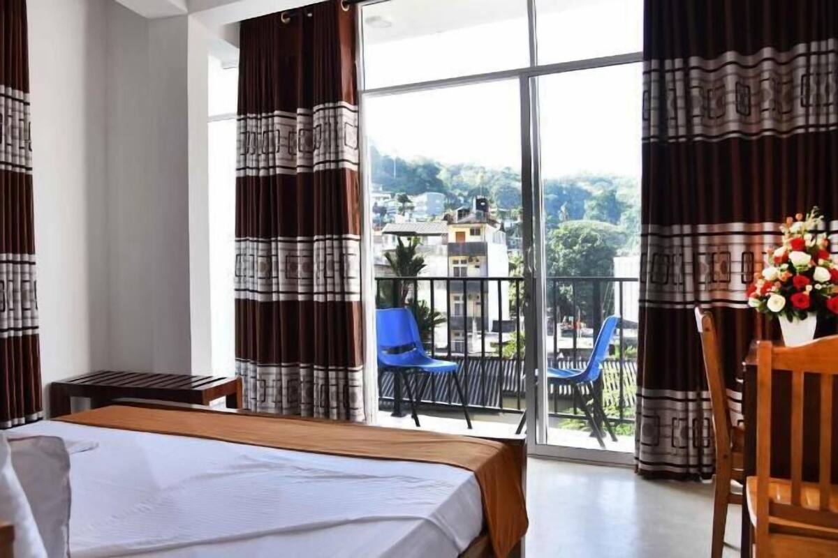 A hotel room with a bed, a small table, a chair, a balcony with a view of buildings and trees.