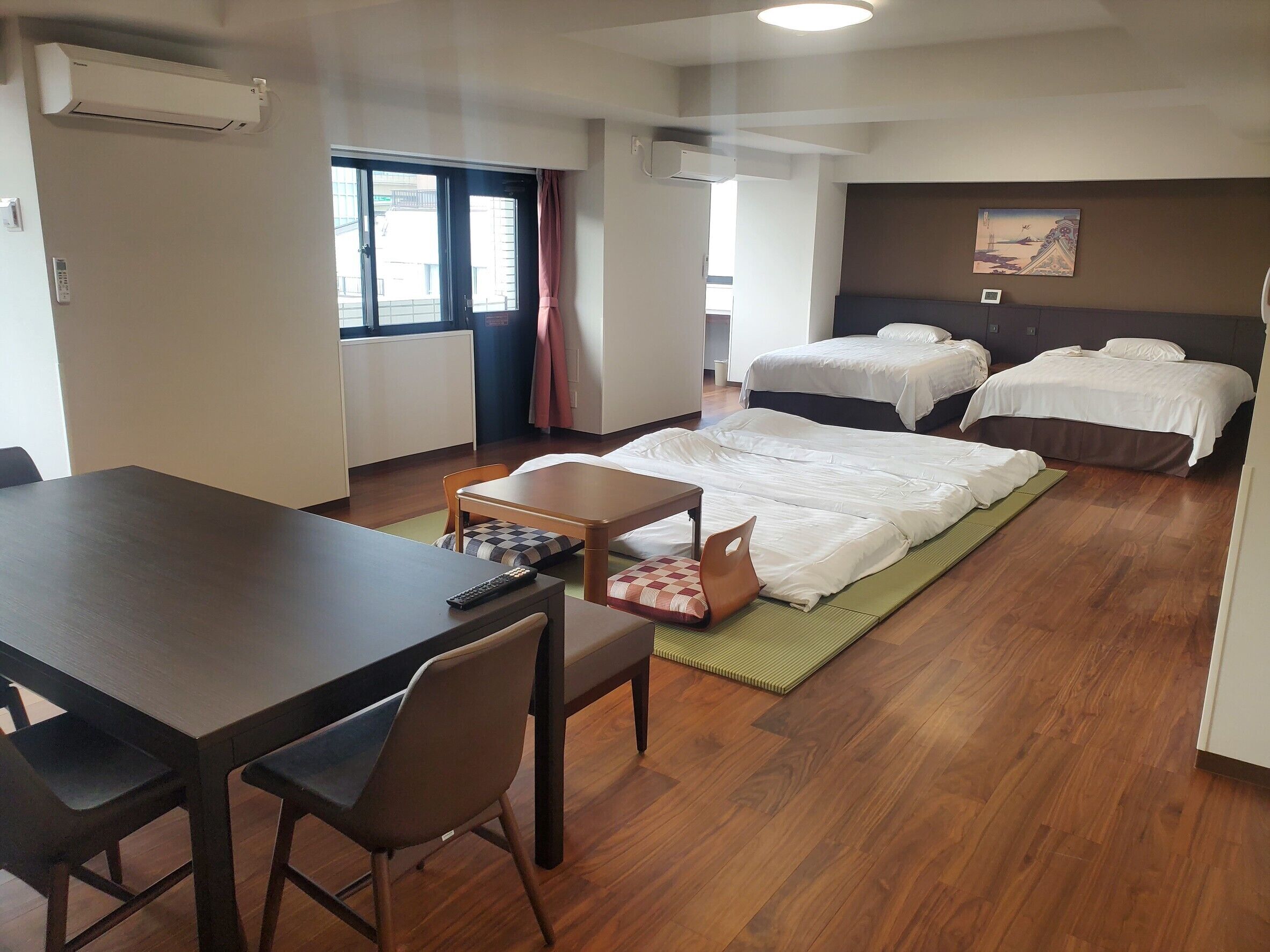 A hotel room with a wooden floor, a desk with a chair, two beds, a window with curtains, and an air conditioning unit.
