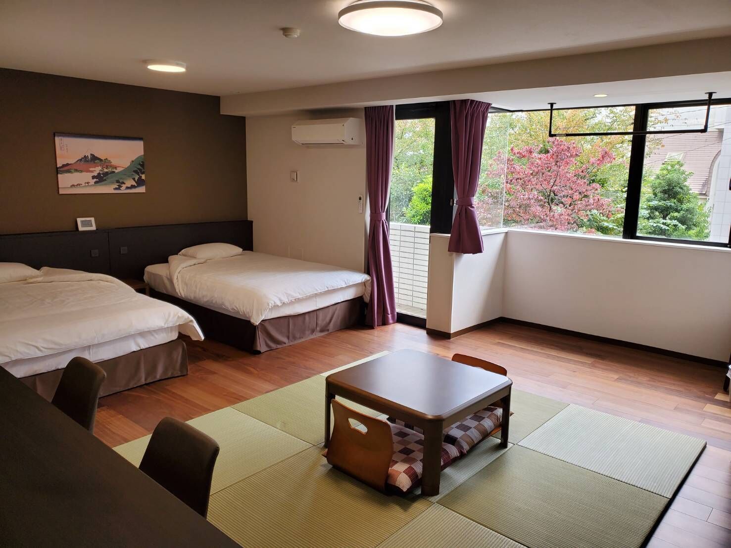 A hotel room with two beds, a dining table, a view of trees and flowers, and a painting on the wall.