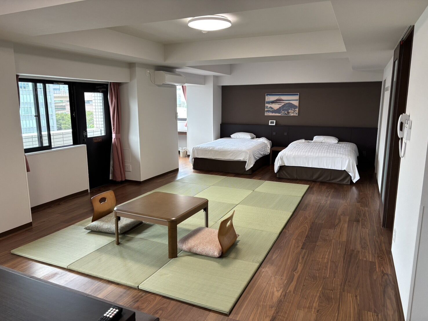 A hotel room with two beds, a desk, a chair, a view of the outside, and a painting on the wall.