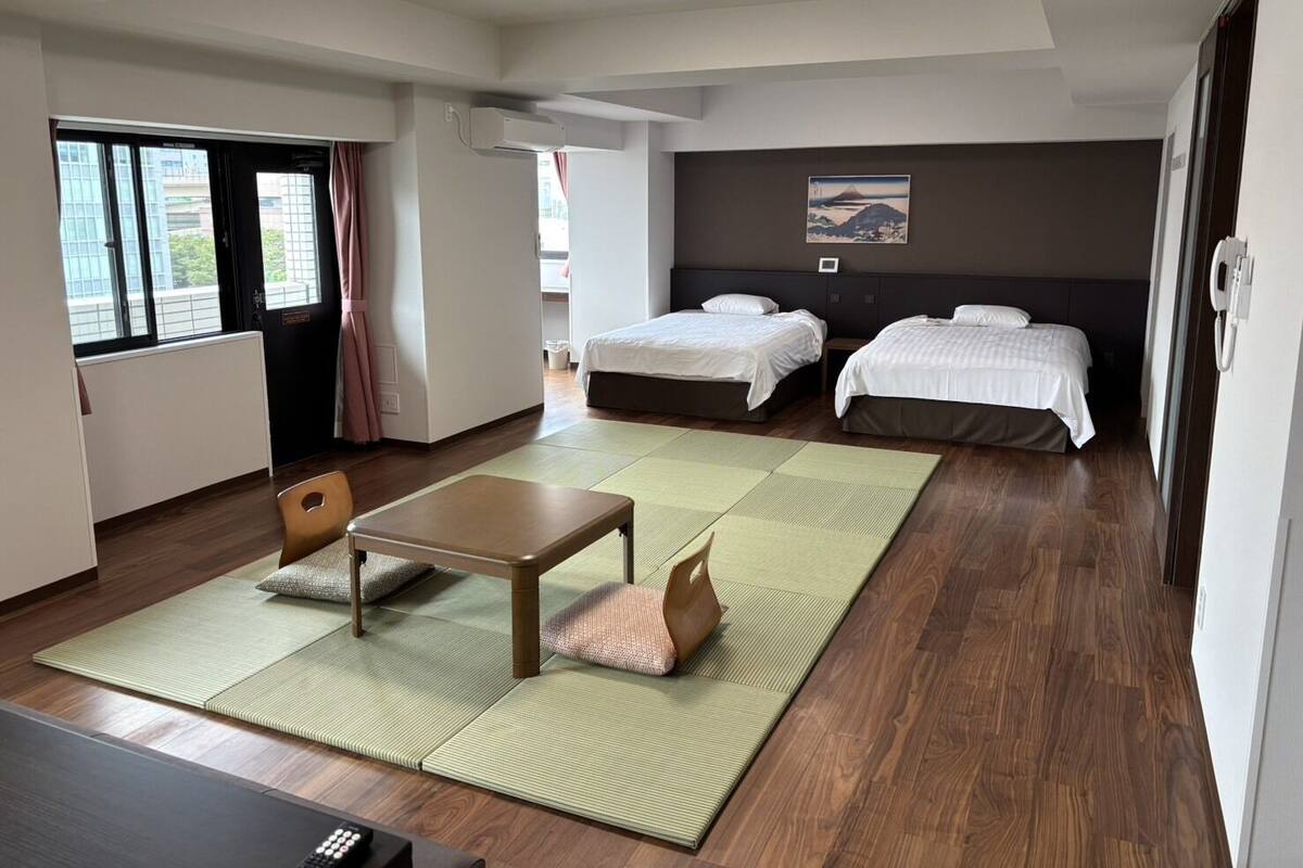 A hotel room with two beds, a desk, a chair, a view of the outside, and a painting on the wall.