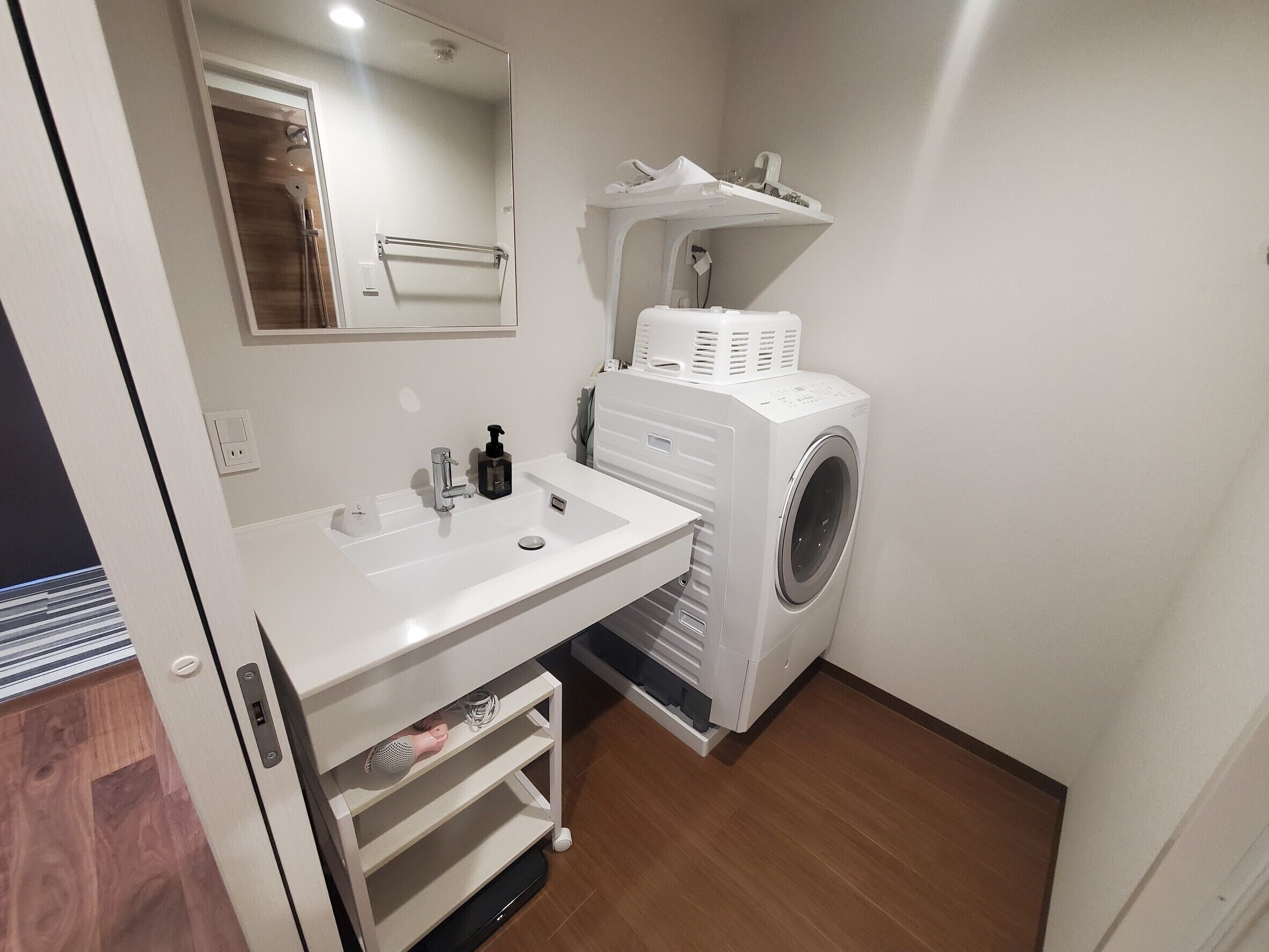 A compact bathroom with a white sink, a mirror, a washing machine, and a shelf with a towel rack.
