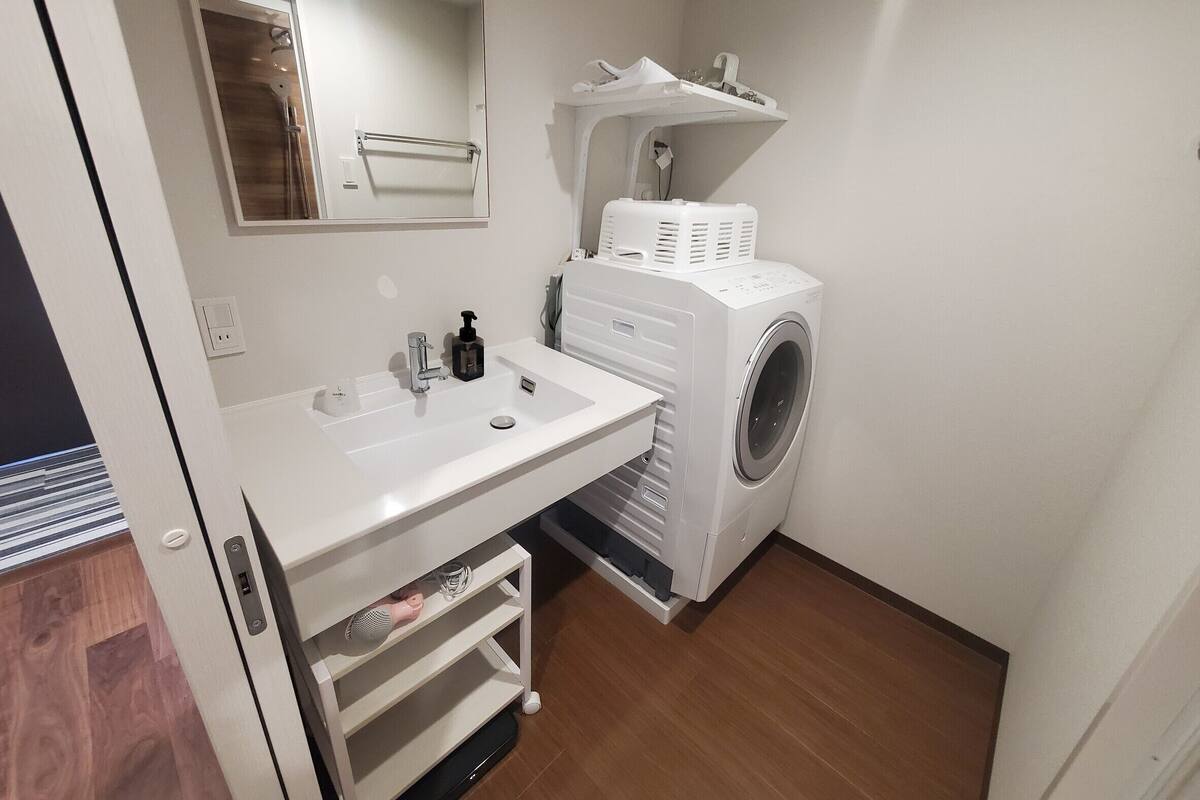 A compact bathroom with a white sink, a mirror, a washing machine, and a shelf with a towel rack.