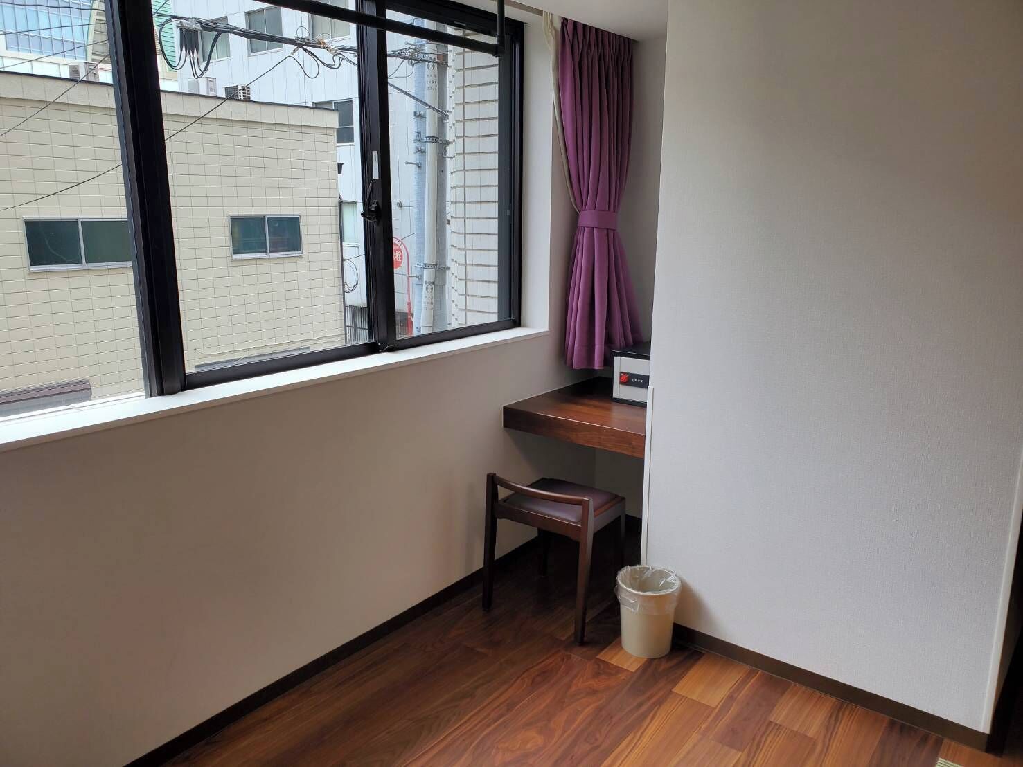 A small room with a wooden floor, a window with a black frame, a purple curtain, a small wooden chair, and a trash bin.