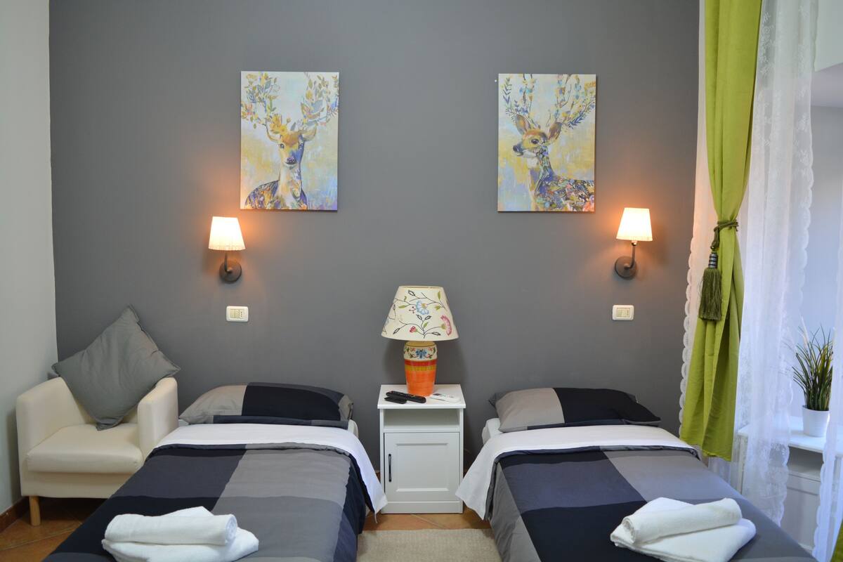 A hotel room with two beds, a nightstand with a lamp, artwork on the wall, and a green curtain.