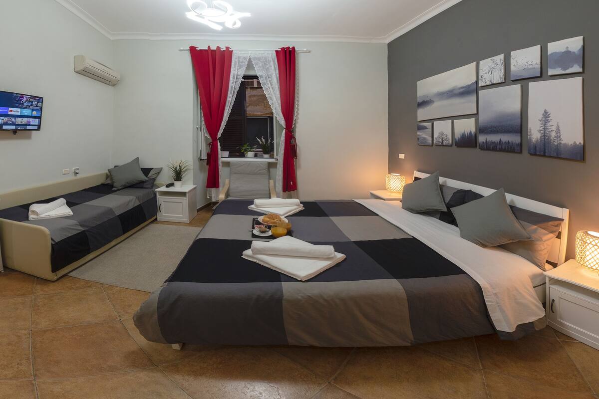 A modern bedroom with a large bed, a sofa, and a desk. There are multiple artworks on the wall and a TV on the wall.