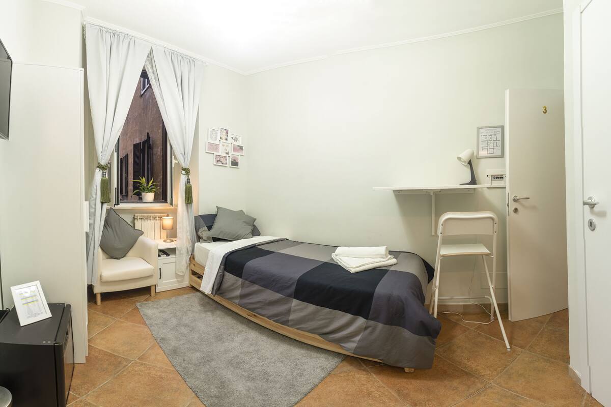 A small, neatly arranged bedroom with a bed, bedside table, chair, and a desk with a chair.