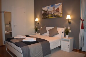 Una cama bien hecha con colcha gris y blanca, dos lámparas de noche, una mesita de noche con una planta y un cuadro de un paisaje urbano en la pared.