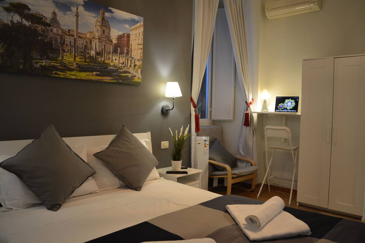 A hotel room with a bed, a desk with a lamp, a chair, a TV, and a wardrobe.