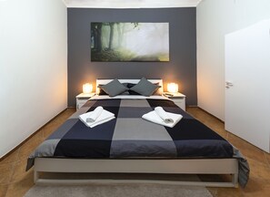 A neatly made bed with a checkered bedspread, two pillows, and two rolled towels. There are two bedside lamps and a painting of a forest on the wall. - Martini Rooms (Rome)