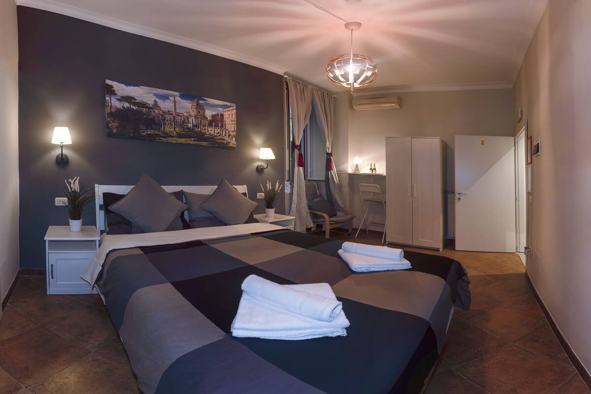 A modern hotel room with a large bed, two bedside tables with lamps, a desk, a chair, a wardrobe, and a painting on the wall.