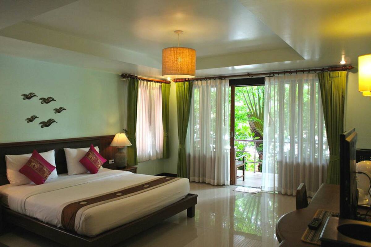 A hotel room with a bed, a desk, a lamp, curtains, and a view of greenery outside.