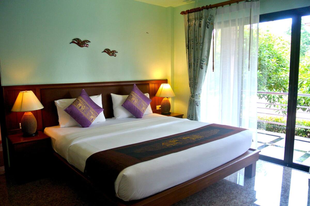 A hotel room with a large bed, two nightstands with lamps, a wooden headboard, and a view of greenery through the window.