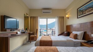 A hotel room with two beds, a desk with a flat-screen TV, a chair, and a balcony with a mountain view.