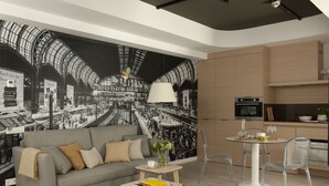 Una sala de estar moderna con un sofá gris, una mesita con un libro y una silla de vidrio transparente. En la pared, un mural grande en blanco y negro de una estación de tren.