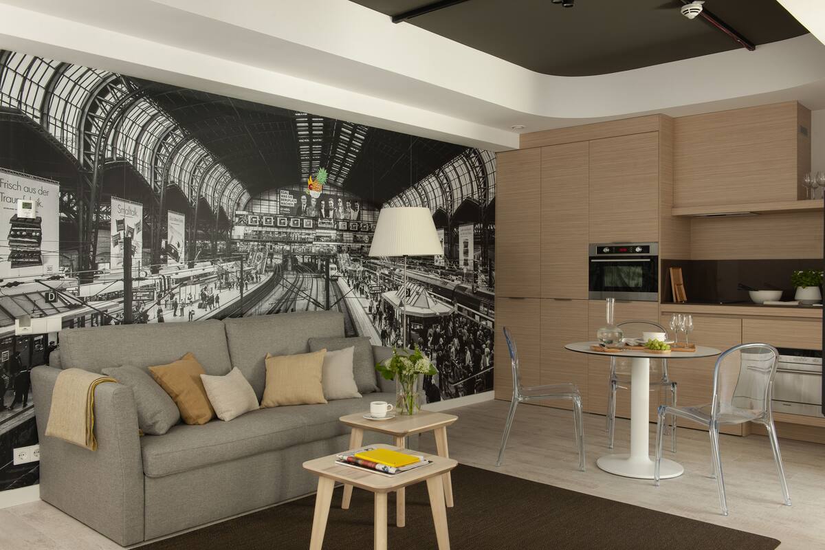 Una sala de estar moderna con un sofá gris, una mesita con un libro y una silla de vidrio transparente. En la pared, un mural grande en blanco y negro de una estación de tren.