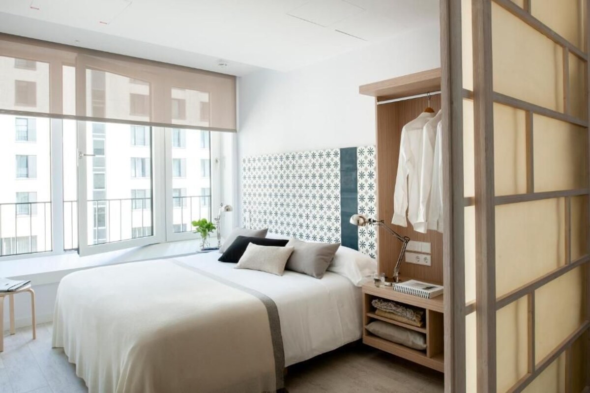 A modern hotel room with a large bed, a bedside table with a lamp, a wardrobe, and a view of the city through large windows.