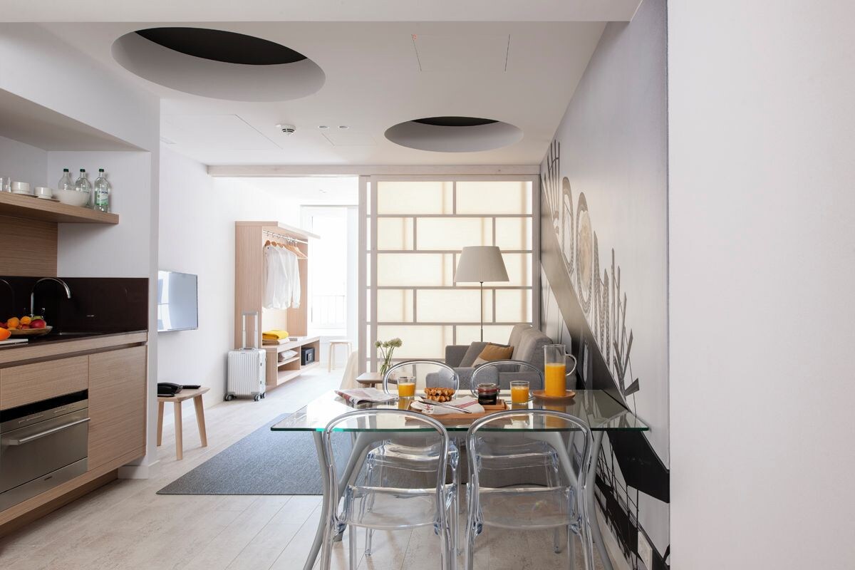 A modern kitchen with a dining area, featuring a glass table and chairs, a sofa, and a large window with natural light.