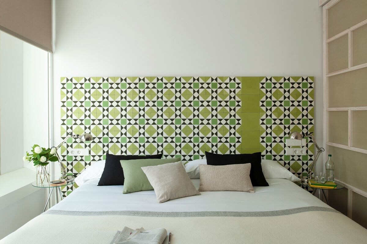 A neatly made bed with a geometric patterned headboard, a bedside table with a lamp, and a window with a view.
