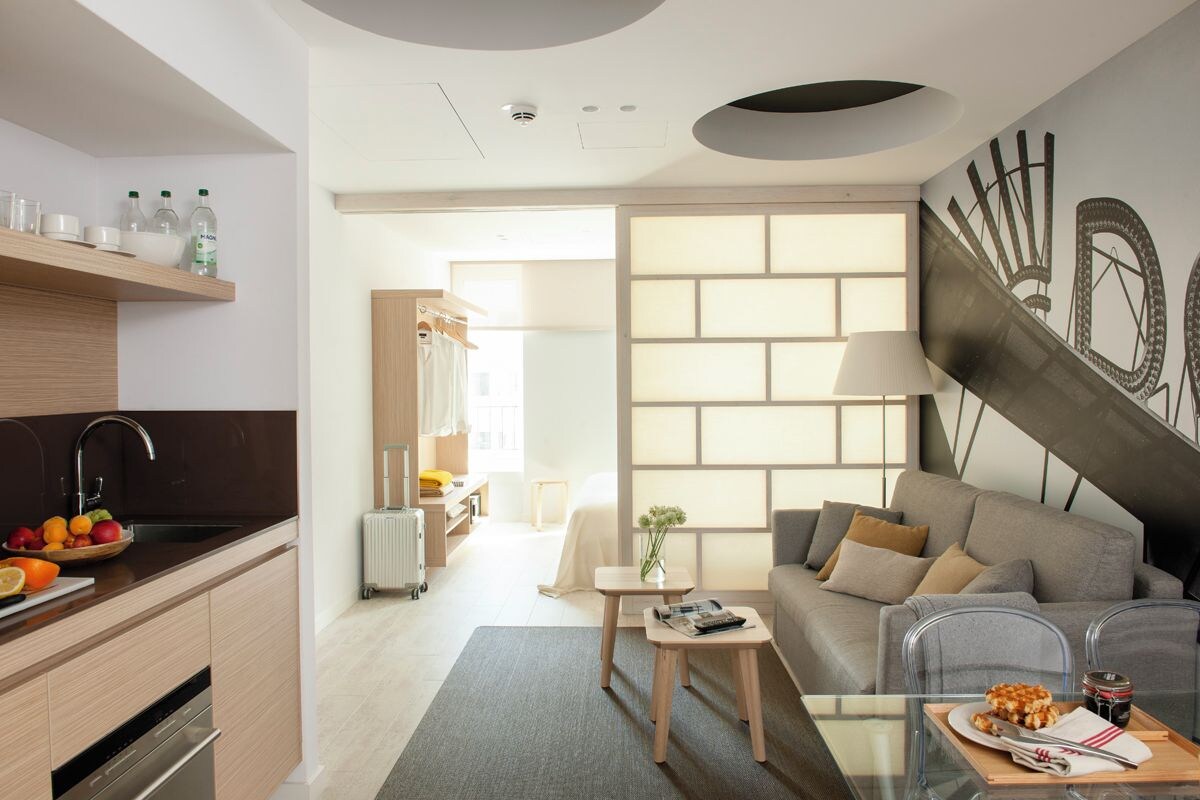 A modern hotel room with a kitchen area, a dining table with a tray of food, a sofa, and a large abstract wall art piece.
