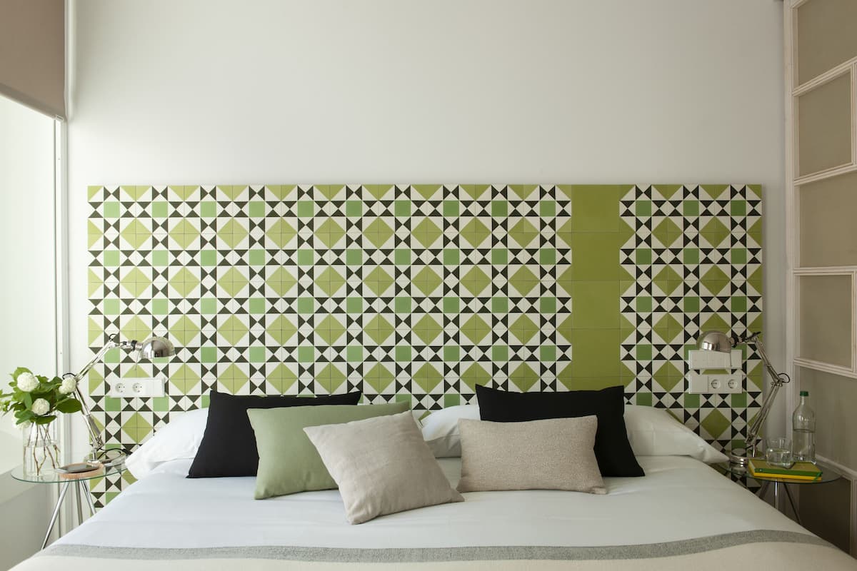 Una cama bien hecha con almohadas, una mesita de noche con lámpara y una pared con un diseño geométrico de azulejos.
