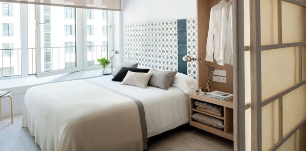 A modern bedroom with a large bed, a bedside table with a lamp, and a view of the city through large windows. - Eric Vökel Boutique Apartments - Hamburg Suites (Hamburg)