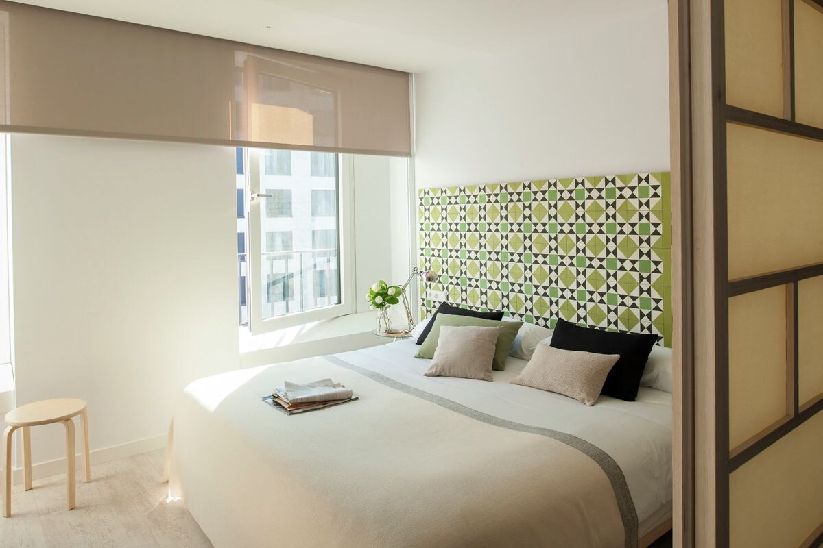 A modern bedroom with a large bed, a window with a view of buildings, a small table, and a patterned headboard.