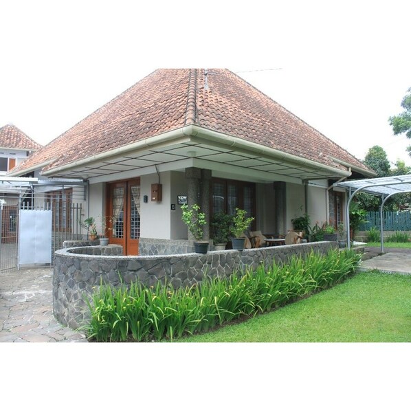 A house with a tiled roof, a stone wall, and a covered porch with potted plants. - De Riau Cottage by HouseinBandung (Bandung)