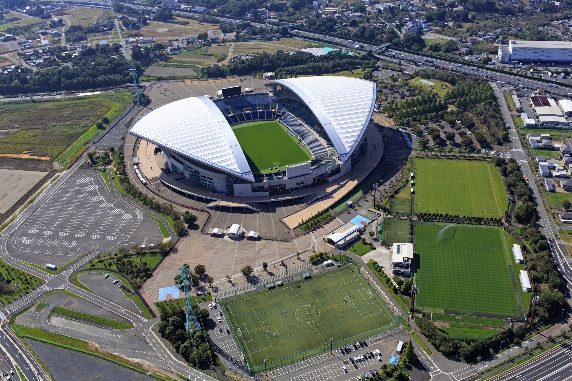 A stadium with a unique curved roof structure, surrounded by parking areas and roads.