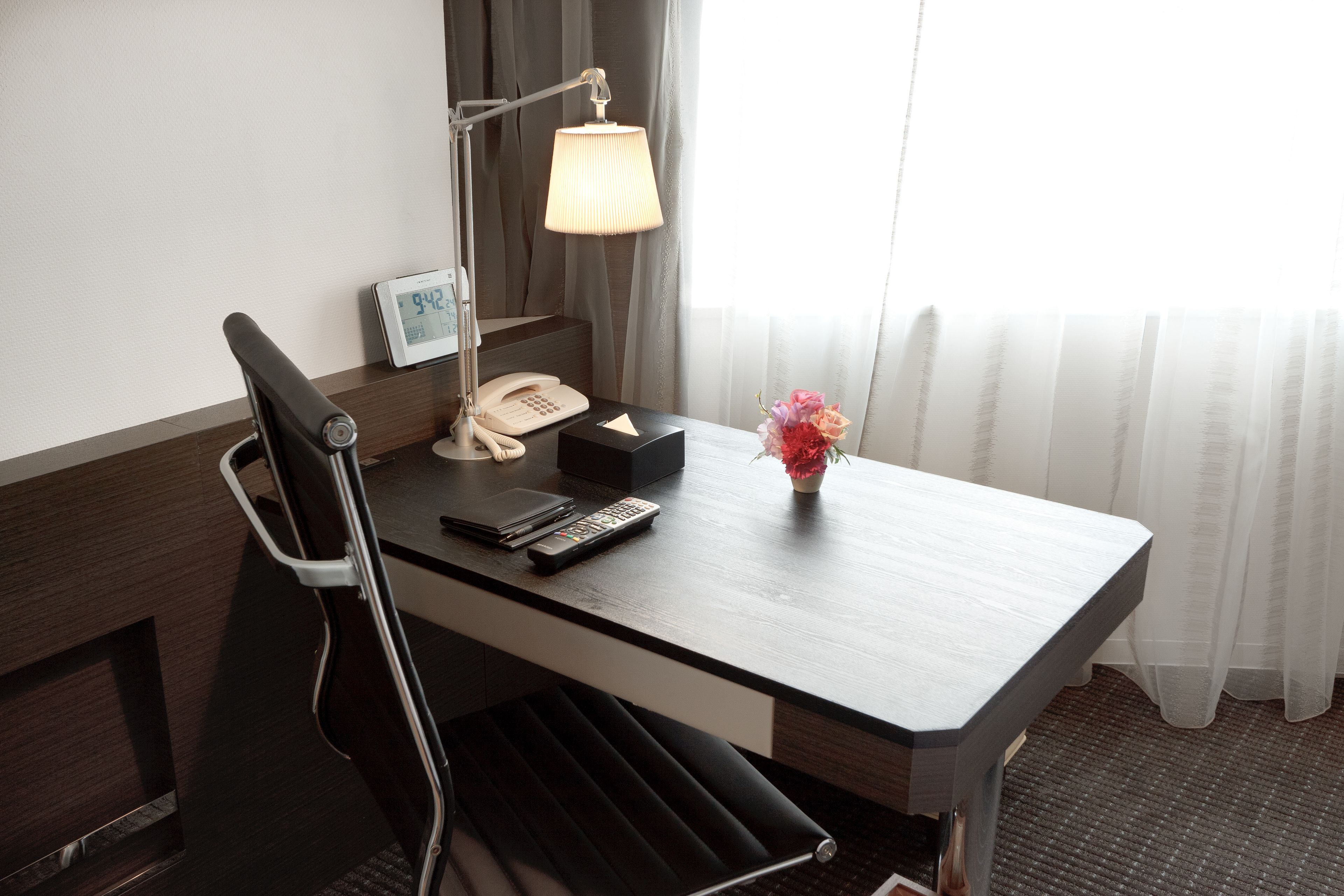 A hotel room with a wooden desk, a lamp, a telephone, a remote control, and a vase with flowers.