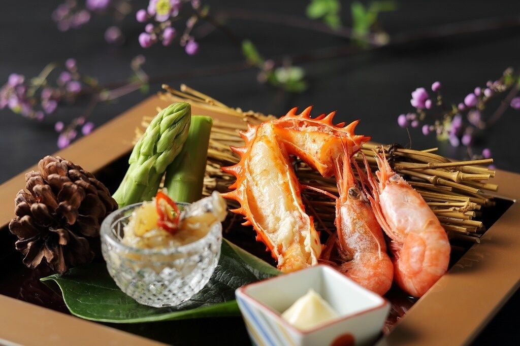 A seafood platter with shrimp, asparagus, and a pine cone on a dark background with purple flowers.