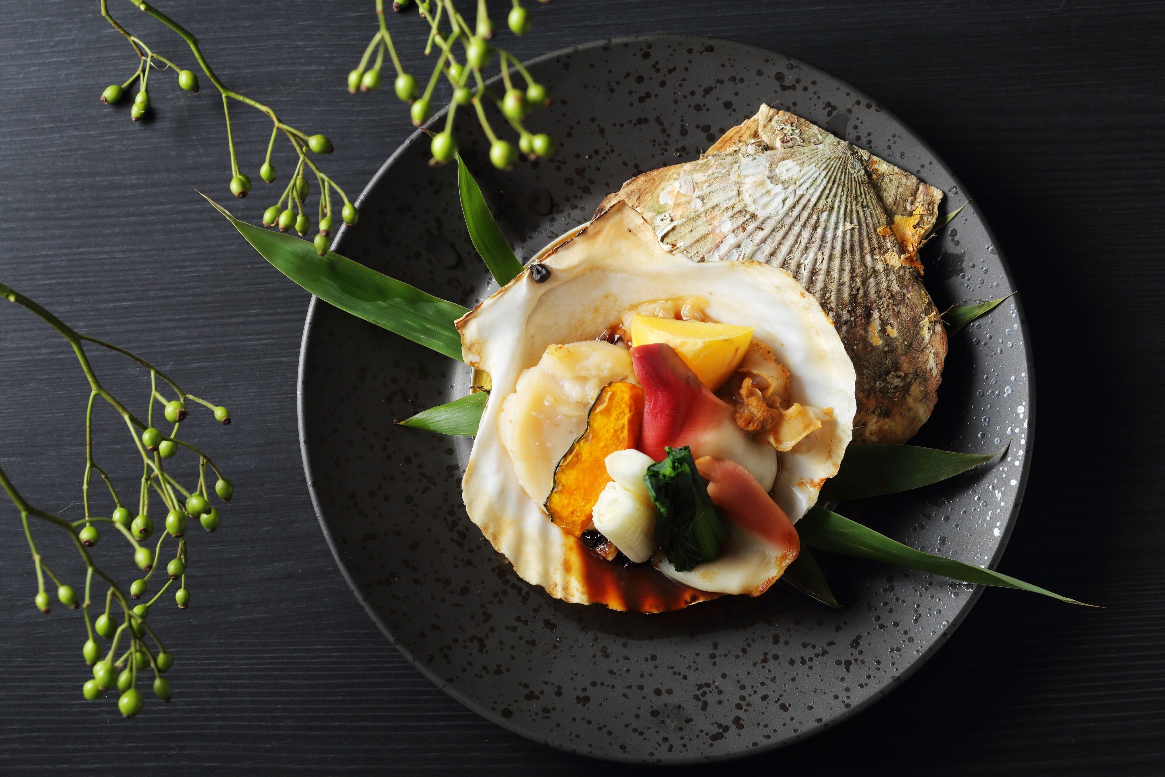 A plate with a scallop, mango, and vegetables on a dark plate.