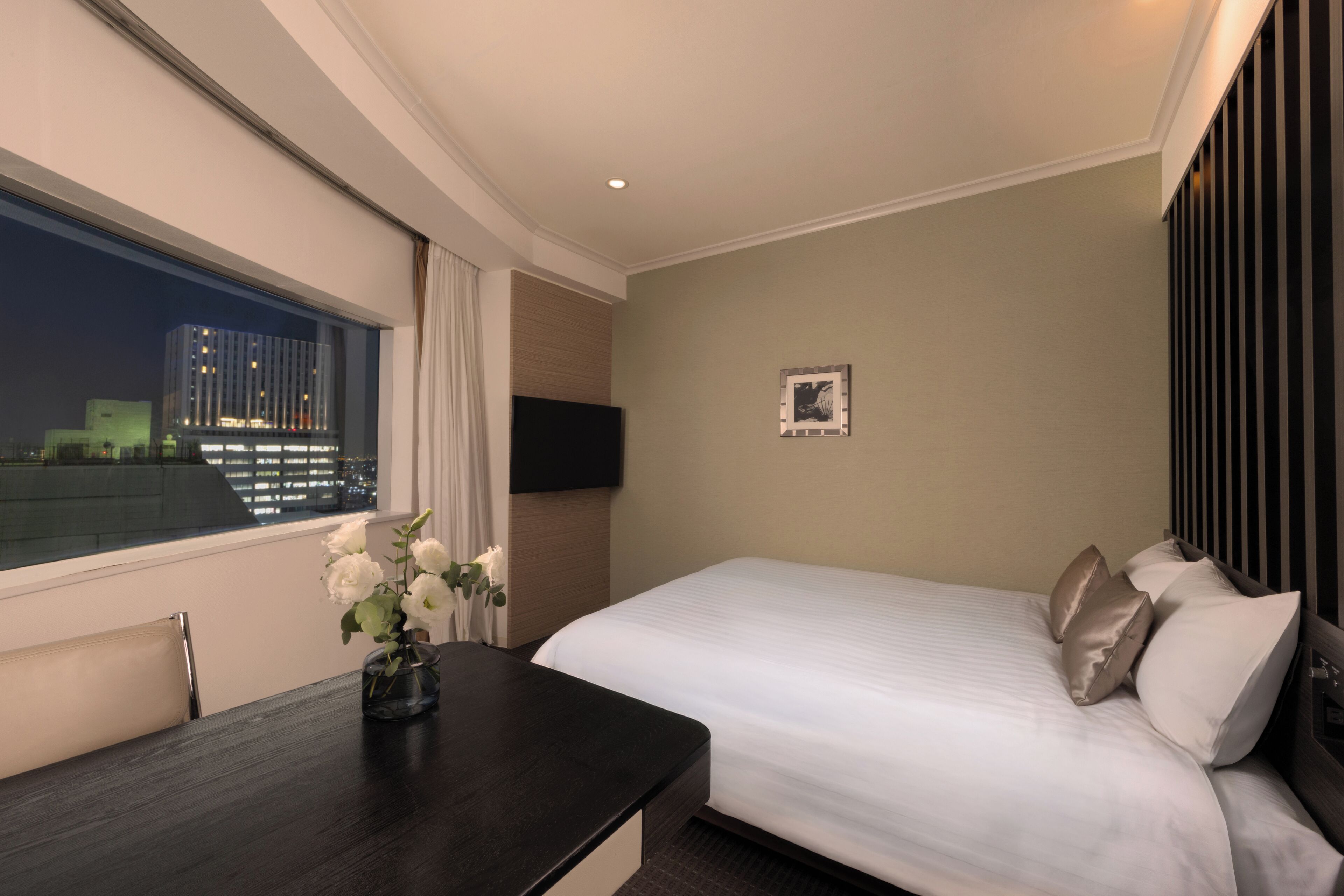 A hotel room with a bed, a desk, a vase with flowers, a flat-screen TV, and a city view.