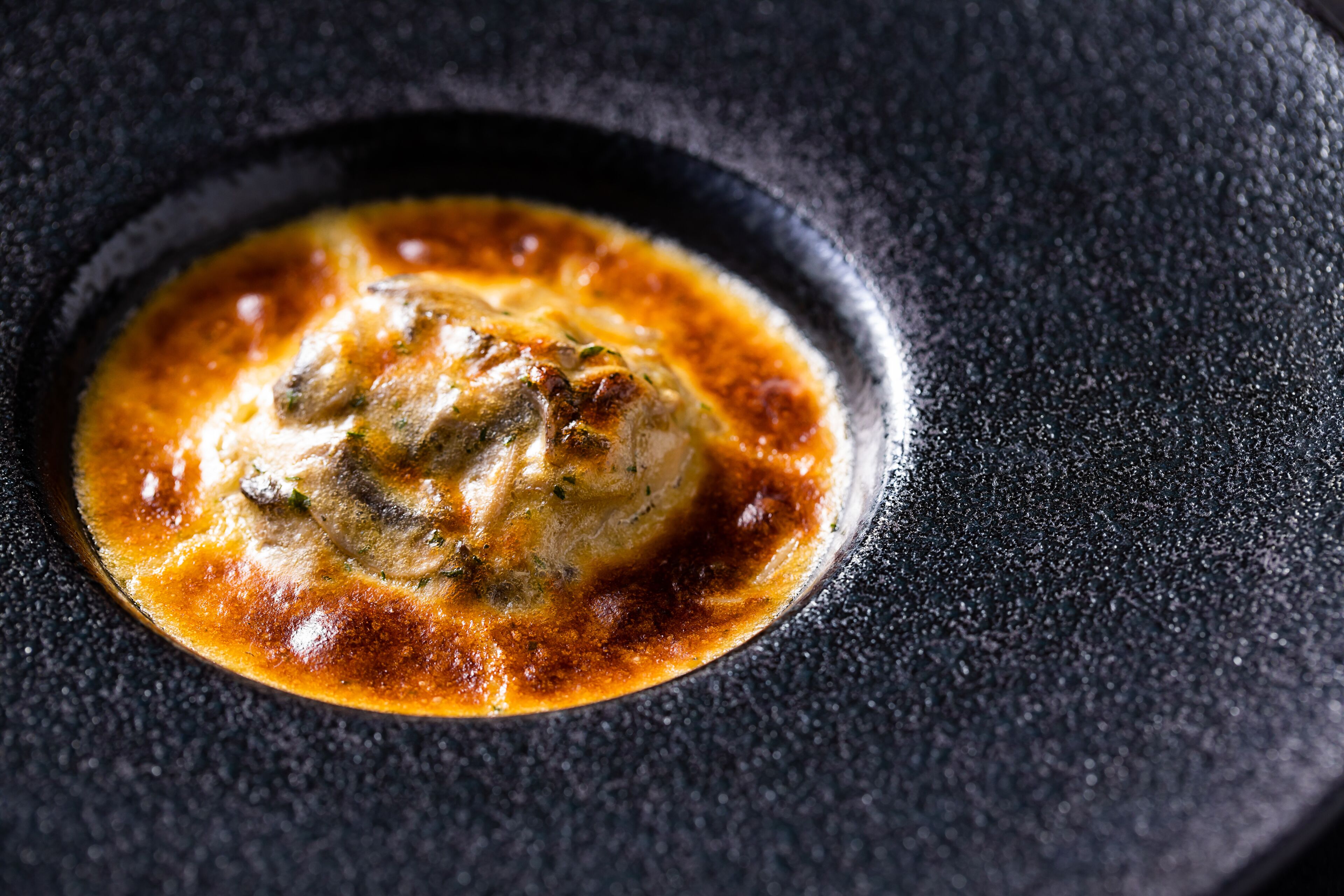Primer plano de un plato con salsa dorada y un bocado en el centro, servido sobre un plato oscuro y con textura.