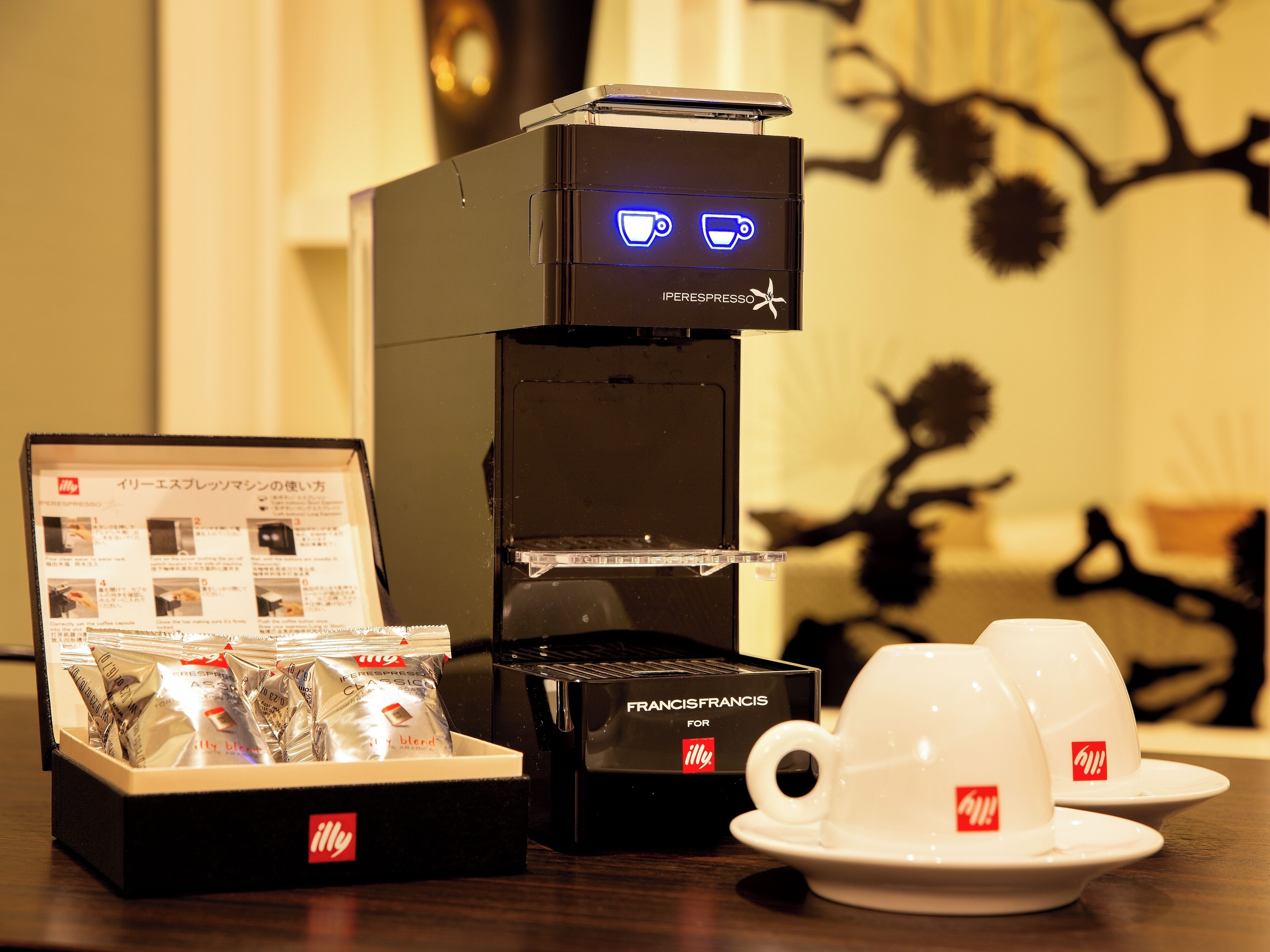 A coffee machine with a digital display, a box of coffee pods, and two white cups with a logo.