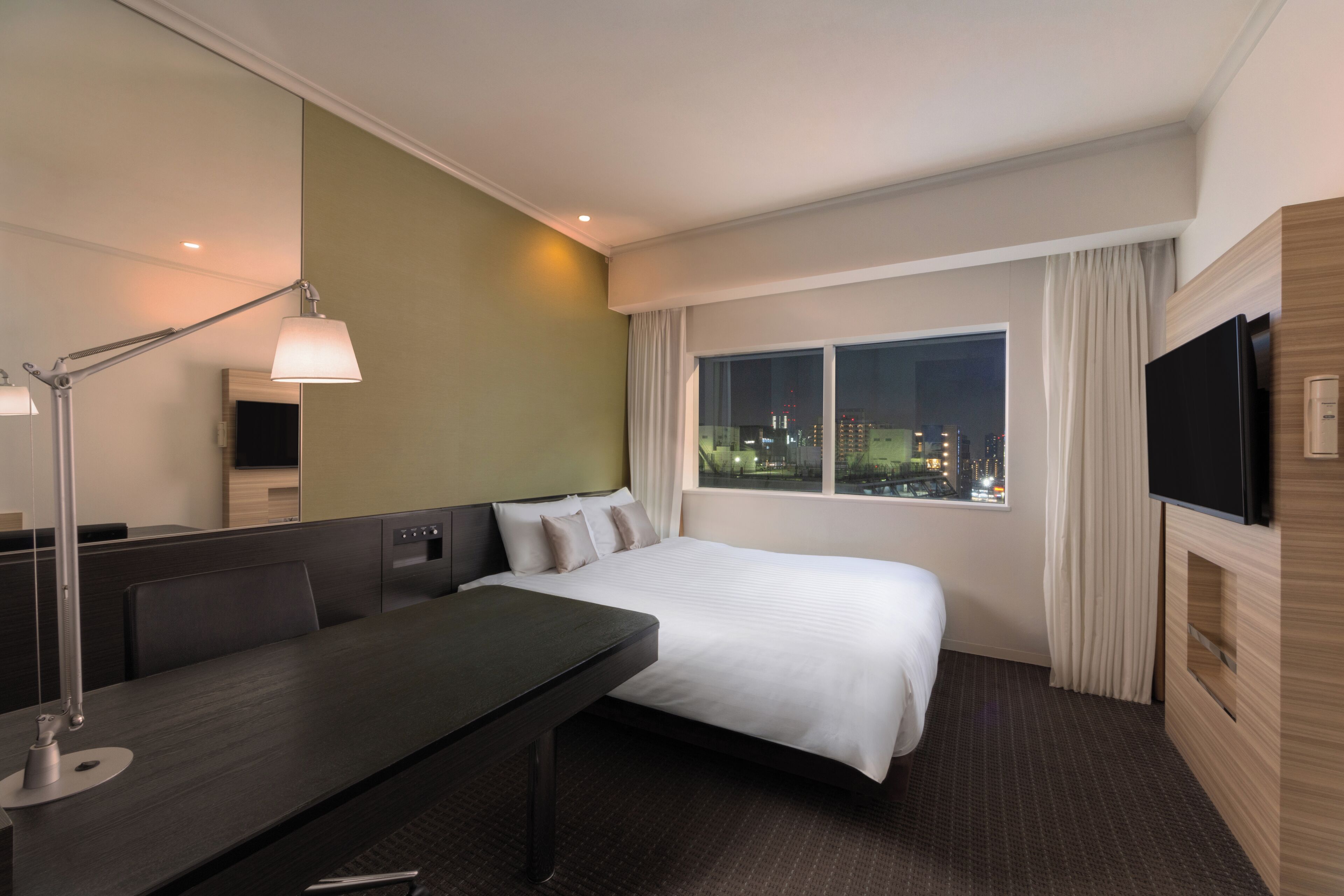 A hotel room with a bed, a desk, a lamp, a TV, and a window with a city view.