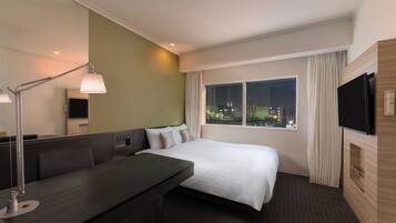 A hotel room with a bed, a desk, a lamp, a TV, and a window with a city view.