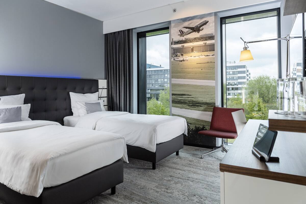 Een hotelkamer met twee bedden, een bureau en een groot raam met uitzicht op een luchthaven en gebouwen.