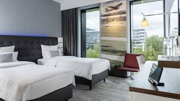 Een hotelkamer met twee bedden, een bureau en een groot raam met uitzicht op een luchthaven en gebouwen.